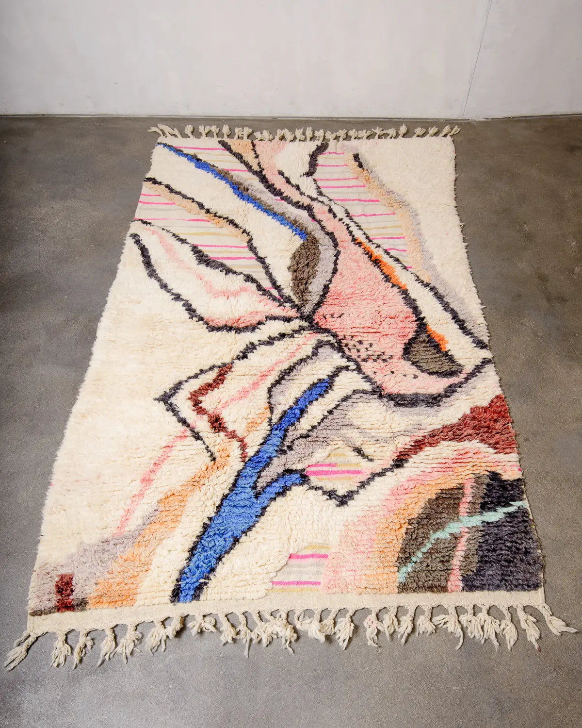 Berberlin Wool Berber rug in shades of cream, blue, pink and orange laying on concrete floor. The rug has a design made of knotted and flat-woven areas.